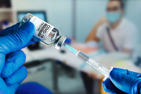 hands of a doctor preparing a syringe to inject the vaccine for immunization to covid-19 in a patient sitting in the medical consultation. Vaccination for covid-19 conceptの写真素材