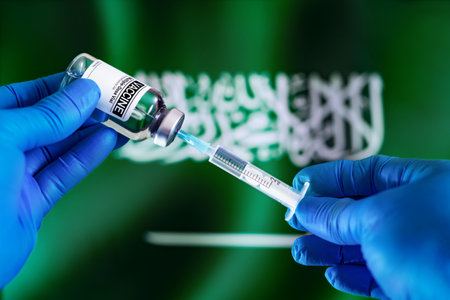 Doctor preparing vial of vaccine injection for the vaccination plan against diseases in Saudi Arabia. Injecting dose of vaccine in syringe for infections prevention in front of the Saudi Arabia flagの写真素材