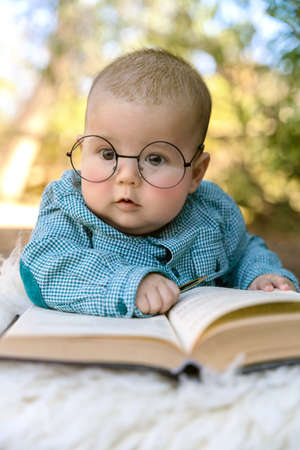 Funny portrait of cute baby in glasses. The baby lies on his stomach and reads an old book on a green background.の写真素材