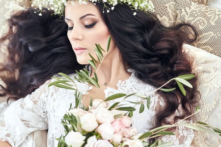 Beautiful bride in lingerie and with a wreath of flowers on her head, in the morning before the wedding. White negligee of the bride, preparing for the wedding ceremony. Sexy girl on the bedの写真素材