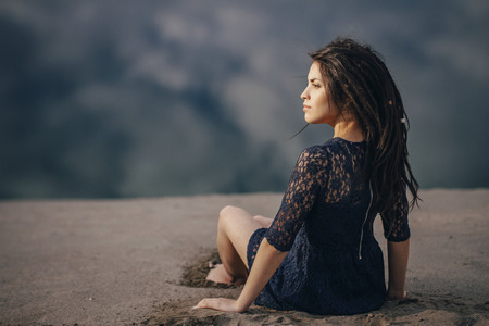 Lifestyle portrait of a woman brunettes in background of lake sitting in sand on a cloudy day. Romantic, gentle, mystical, pensive image of a girl. Girl Oriental appearance, dreams of sitting on sandの写真素材