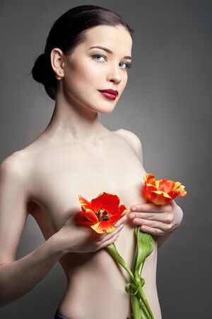 Nude Naked girl with tulips flowers in hand and perfect skin, sexy naked brunette woman to gray background. Skin care of face and hands in the summer. Bright natural makeupの写真素材
