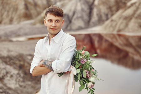 Man in a white shirt and a bouquet of flowers in his hands waiting for a girl on a date. Portrait of a man with flowers on the background of beautiful mountainsの写真素材