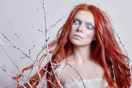 Redhead girl with long hair, a face covered with snow with frost. White eyebrows and eyelashes in frost, a tree branch covered with snow. Snow Queen and winter. Winter makeup woman face, red headの写真素材