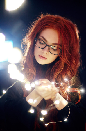 Art portrait of a woman with red hair in Christmas lights. Girl in glasses with reflected Christmas...
