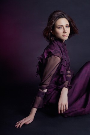 Contrast fashion Armenian woman portrait with big blue eyes sit on a dark background in a purple dress. Lovely gorgeous girl posing in evening dress. Bright makeup, womanの写真素材