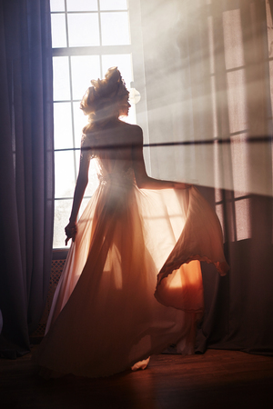Silhouette of a sexy woman girl near the window in the morning evening sun. Fashion blonde woman in the rays of sun. Sunlight falls on a woman's body. Fairy magic light in windowの写真素材