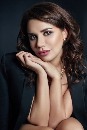 Sexy brunette with beautiful curly hair in an erotic black jacket. Expensive jewelry and smooth natural pure skin. Beautiful evening makeup, fashion portrait of a womanの写真素材