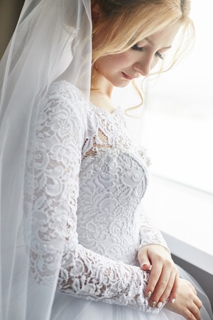 Portrait of a bride in a chic white wedding dress preparing for the wedding ceremony. Portrait of a blonde woman near a window in a white dress and a veil on her head. Bride is waiting for her groomの写真素材