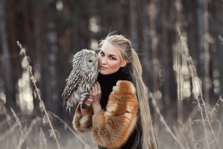 Woman blond in autumn in fur coat with owl on hand first snow. Beautiful girl with long hair in nature, holding an owl. Romantic, delicate look girlsの写真素材