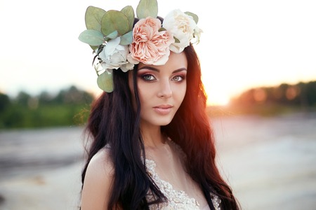 Woman with a wreath of flowers on her head and a summer dress walks through hills at sunset. Portrait of a brunette with long hair in rays of the setting sun, perfect makeup and beautiful blue eyesの写真素材