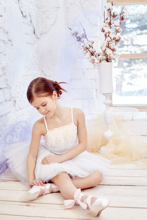 Little prima ballet. Young ballerina girl is preparing for a ballet performance. Girl in a white ball gown and Pointe near the window, beautiful red hair. Young theater actressの写真素材