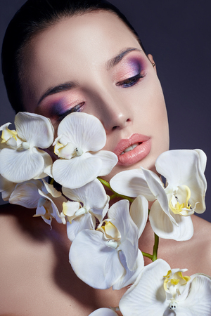 Woman with white Orchid near face girl with beautiful bright makeup pink lipstick. Gentle art portrait Advertising cosmetics, professional makeup art, glitter lips, bright color, blue shadows on eyesの写真素材