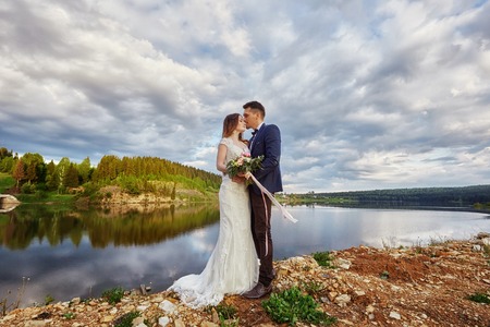 Beautiful couple in love kissing while standing on ground by lake. Wedding couple at sunset, blue cloud sky, love and tender feelings. Loving couple resting. Wedding ceremony outdoors. Perfect coupleの写真素材