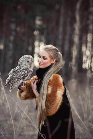 Woman blond in autumn in fur coat with owl on hand first snow. Beautiful girl with long hair in nature, holding an owl. Romantic, delicate look girlsの写真素材
