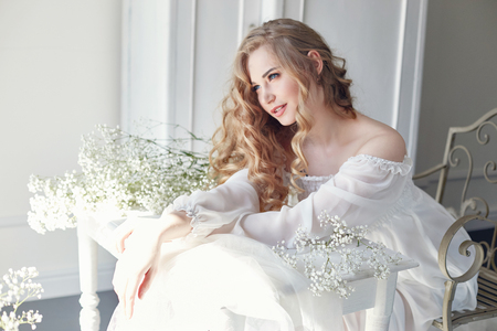 Girl white light dress and curly hair, portrait of woman with flowers at home near the window, purity and innocence. Curly blonde romantic look, beautiful eyes. White wildflowers in handsの写真素材