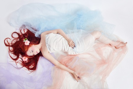 Redhead girl in light airy colored dress lies on the floor white background. Beautiful flowers in girl hair. Romantic woman with long hair and cloud dress. Girl dreaming bright makeup and perfect bodyの写真素材