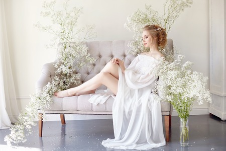 Girl writes a letter to her beloved man, sitting at home at table in a white light dress, purity and innocence. Curly blonde romantic look, beautiful eyes. White wildflowers on the table. Perfect bodyの写真素材