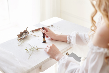 Girl writes a letter to her beloved man, sitting at home at table in a white light dress, purity and innocence. Curly blonde romantic look, beautiful eyes. White wildflowers on the table. Perfect bodyの写真素材