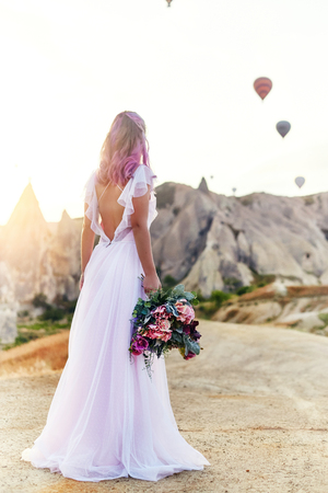 Woman in a long dress on background of balloons in Cappadocia. Girl with flowers hands stands on a hill and looks at a large number of flying balloons. Turkey Cappadocia fairytale scenery of mountainsの写真素材