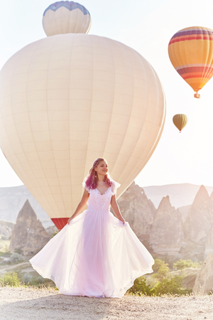 Woman in a long dress on background of balloons in Cappadocia. Girl with flowers hands stands on a hill and looks at a large number of flying balloons. Turkey Cappadocia fairytale scenery of mountainsの写真素材