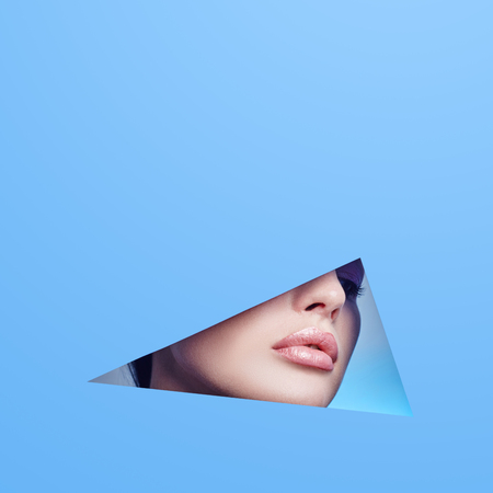 Woman looking in the hole, bright beautiful makeup, big eyes and lips, bright lipstick, professional cosmetics and facial care. Bright colored background and a gap slot in the paperの写真素材