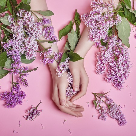 Fashion art hands natural cosmetics women, bright purple lilac flowers in hand with bright contrast makeup, hand care. Creative beauty photo of a girl sitting at table on contrasting pink backgroundの写真素材