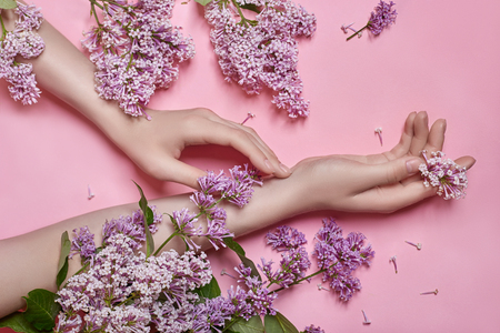 Fashion art hands natural cosmetics women, bright purple lilac flowers in hand with bright contrast makeup, hand care. Creative beauty photo of a girl sitting at table on contrasting pink backgroundの写真素材