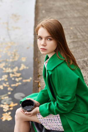 Girl in a green coat walks along the lake embankment on a cloudy autumn day. Autumn fashion and clothing, yellow fallen leaves floating in the water. Romantic moodの写真素材