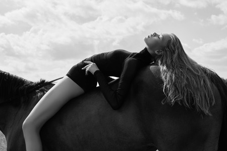 Girl rider lies bent on a horse in the field. Fashion portrait of a woman and the mares are horses in the village in the sky. Blonde woman lies and dreams on a horse, beautiful girl bodyの写真素材