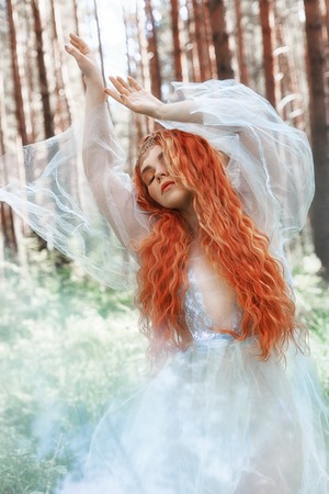 Beautiful redhead woman forest nymph in a blue transparent light dress in the woods spinning in dance. Red hair girls. Art fashion portrait of fairy woman fairy tale in summer forestの写真素材