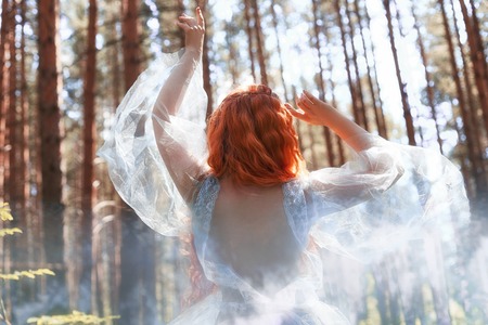 Beautiful redhead woman forest nymph in a blue transparent light dress in the woods spinning in dance. Red hair girls. Art fashion portrait of fairy woman fairy tale in summer forestの写真素材
