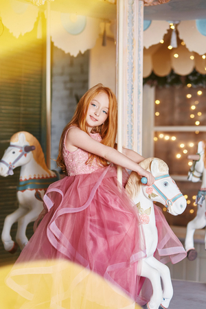 Beautiful red-haired girl with long hair and blue eyes rides a carousel in a long pink dress. Carousel in the form of horses, redhead girl celebrates her birthday in the amusement Parkの写真素材