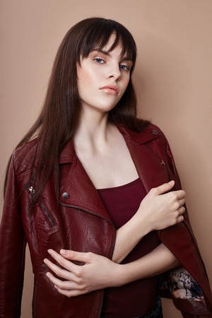 Sexy brunette in leather jacket and red top, portrait bright makeup and lipstick close-up, face makeup, professional makeup, skin care. Young sexy woman posing on beige background, flying hairの写真素材