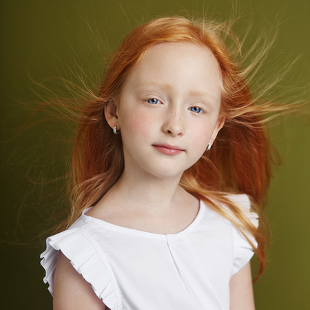 Beautiful red-haired girl with long hair and beautiful big blue eyes. Redhead Girl child in summer clothes posing on the background of olive color. Norwegian girl with bright red hairの写真素材