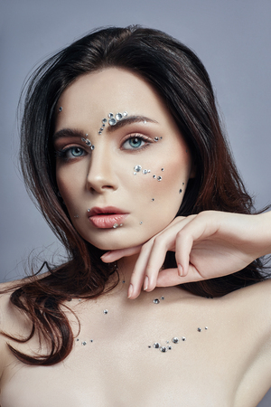 Art portrait of a woman with large crystals of rhinestones on her face, chest and body. Perfect makeup, skin care, professional cosmetics. Girl's face with diamonds on the skin close-upの写真素材