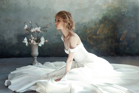 Girl in a chic long dress sitting on the floor. White wedding dress on the bride's body. Beautiful light dress with a long hem, a portrait of a fragile delicate girl before the wedding ceremonyの写真素材