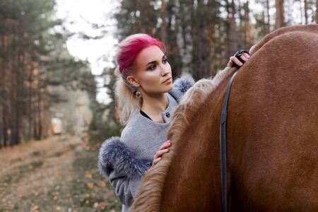 Woman walking with horse autumn on nature. Creative hot pink makeup girl face, hair coloring. Portrait of a girl with a horse. Horse riding in the autumn forest. Autumn clothes and bright red makeupの写真素材