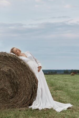 Pregnant girl walks in a field near haystacks in a long white dress, a woman smiles and holds her hands over her stomach. Girl is expecting the birth of a baby in the ninth month of pregnancyの写真素材