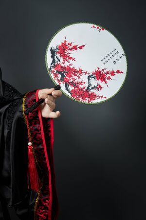 Woman is dressed in red Chinese Japanese folk clothing. Flying fabric, beautiful umbrella and fan in Japanese Chinese style, long earrings in the ears. Girl posing on a dark backgroundの写真素材