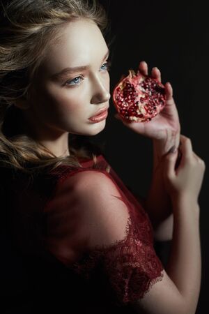 Beautiful blonde girl with pomegranate fruit in her hands. Spring portrait of a girl in a red dress breaking a pomegranate, juice flowing down her handsの写真素材