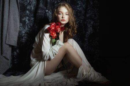Beautiful spring girl is sitting on the floor with rose flowers in her hands. Woman in a white dress is dreaming, a romantic image. Blonde with curly hairの写真素材