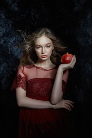 Beautiful blonde girl with pomegranate fruit in her hands. Spring portrait of a girl in a red dress breaking a pomegranate, juice flowing down her handsの写真素材