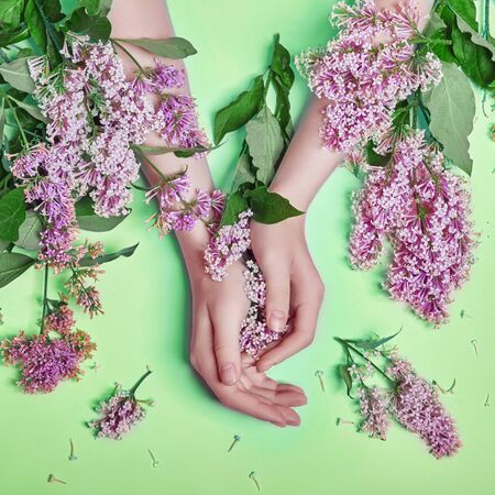 Fashion art hands natural cosmetics women, bright purple lilac flowers in hand with bright contrast makeup, hand care. Creative beauty photo of a girl sitting at table on contrasting green backgroundの写真素材