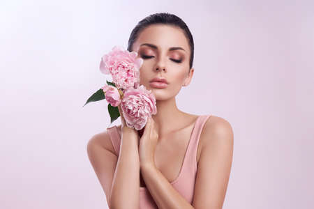 Beautiful woman with pink peony flowers, natural beauty, skin care. Perfect facial features. Girl posing on a light backgroundの写真素材