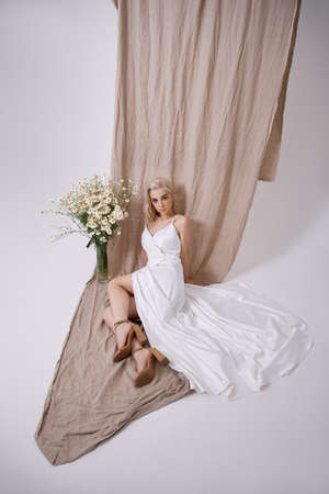 blonde woman in a beautiful white dress is sitting near the window in front of a bouquet of wild flowers. Romantic girl with beautiful natural makeupの写真素材