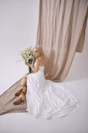 Portrait of a sexy blonde in a beautiful white dress near a vase of wild flowers. Romantic girl with beautiful natural makeupの写真素材