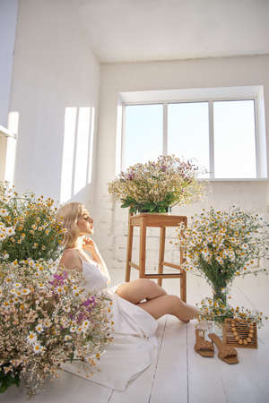Beautiful blonde woman in a white dress is sitting on the floor among the chamomile flowers. Portrait of a girl with a bouquet of wild flowersの写真素材