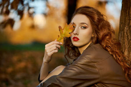 Fashionable redhead woman in a maroon beret and leather raincoat stands on the porch in autumn. Beauty portrait of a woman with red hairの写真素材