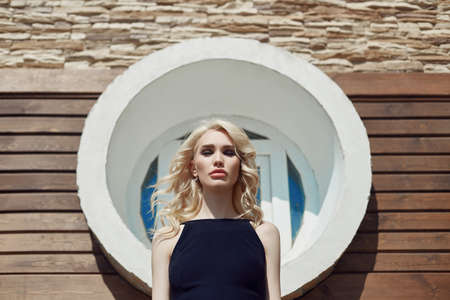 Beautiful portrait of a blonde woman against a brick wall on the street in a black tight dress. Perfect hair styling, curly hair hairstyleの写真素材